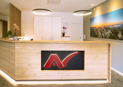 Dr. med. Andreas Nour-El-Din Praxis: Empfangsbereich mit Holztheke, Logo und Bergpanorama-Bild